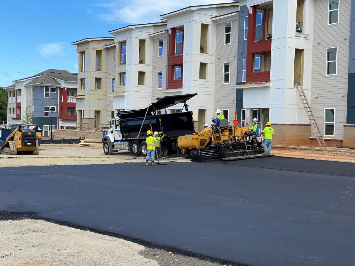Construction crew paving a street in a residential area with a dump truck and asphalt paver nearby.