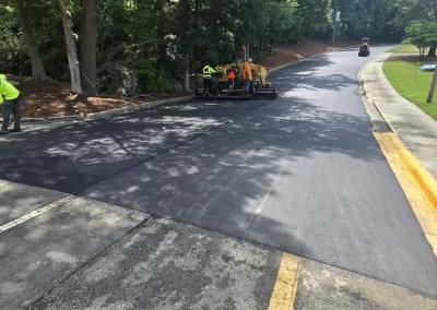Road crews in high-visibility vests paving a curb-to-curb asphalt road, with a yellow paving machine and a vehicle in the distance near a green, tree-lined sidewalk.