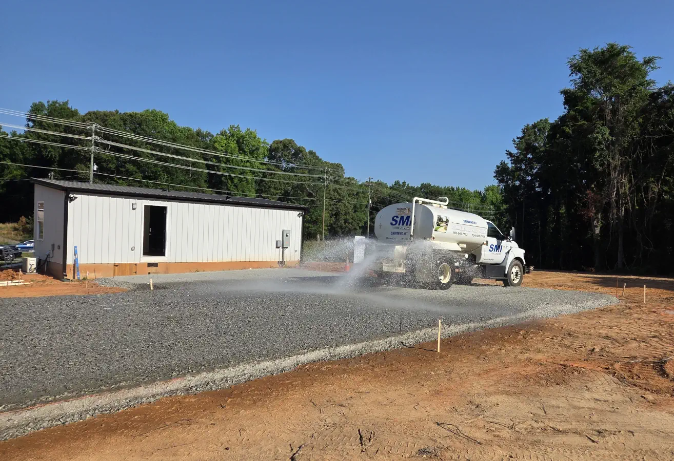 What grading involves Water tanker spraying water onto a gravel construction area, with a white building and trees in the background.